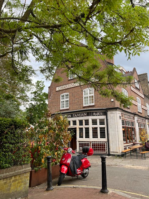 A red scooter parked on the sidewalk in front of a traditional brick pub building with large front windows displaying signage for 'Barton Tavern Ales' and 'Charrington.' The pub has a corner entrance, and outdoor wooden benches are placed nearby. Overhanging green tree branches partially cover the view of the building, which is situated on a street likely in Chiswick. The pavement is edged with black metal bollards, and the scene is captured in natural daylight, illustrating a typical setting for house removals or furniture transport to or from a residential or pub area in Chiswick, with surroundings suitable for moving services by Man with Van Chiswick.