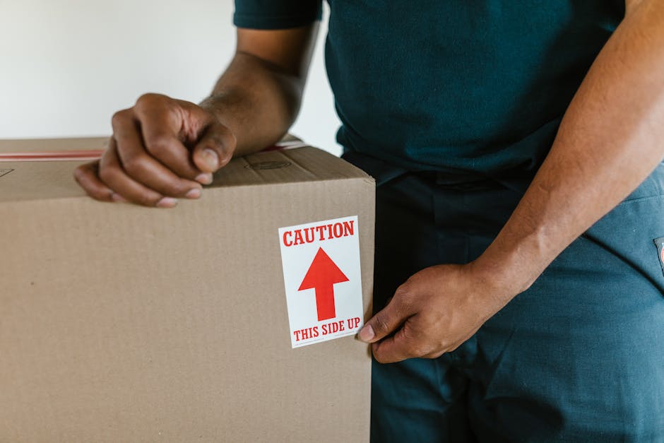Close-up of a person during a home relocation process, wearing a dark green T-shirt and dark trousers, while holding a cardboard box with both hands. The box features a prominent red and white label w