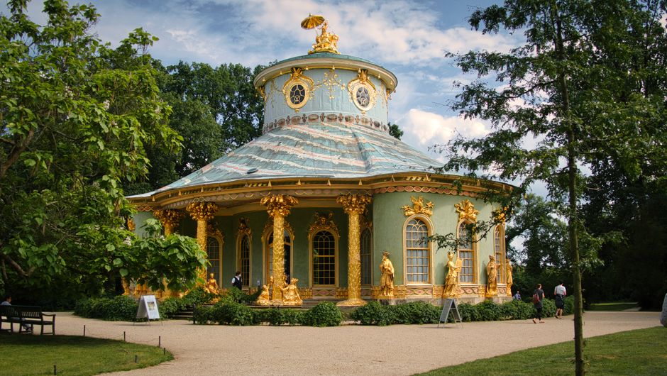A large, white, neoclassical-style mansion with multiple columns and decorative balustrades on the upper terrace is situated behind a well-maintained lawn and garden. In the foreground, a calm body of