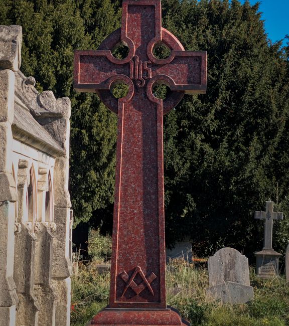 A tall red granite Celtic cross grave marker situated outdoors in a cemetery, surrounded by green trees and several other headstones, including a weathered stone monument on the left and smaller gray headstones in the background. The cross features intricate circular and linear carvings, with a prominent Masonic square and compass symbol near its base. The ground around the monument is covered with grass and wild plants, and the scene is lit by natural daylight, highlighting the texture and color of the stone. This setting suggests a peaceful, well-maintained graveyard environment, consistent with house removals and relocations involving careful handling of memorial monuments, as managed by Man with Van Chiswick.